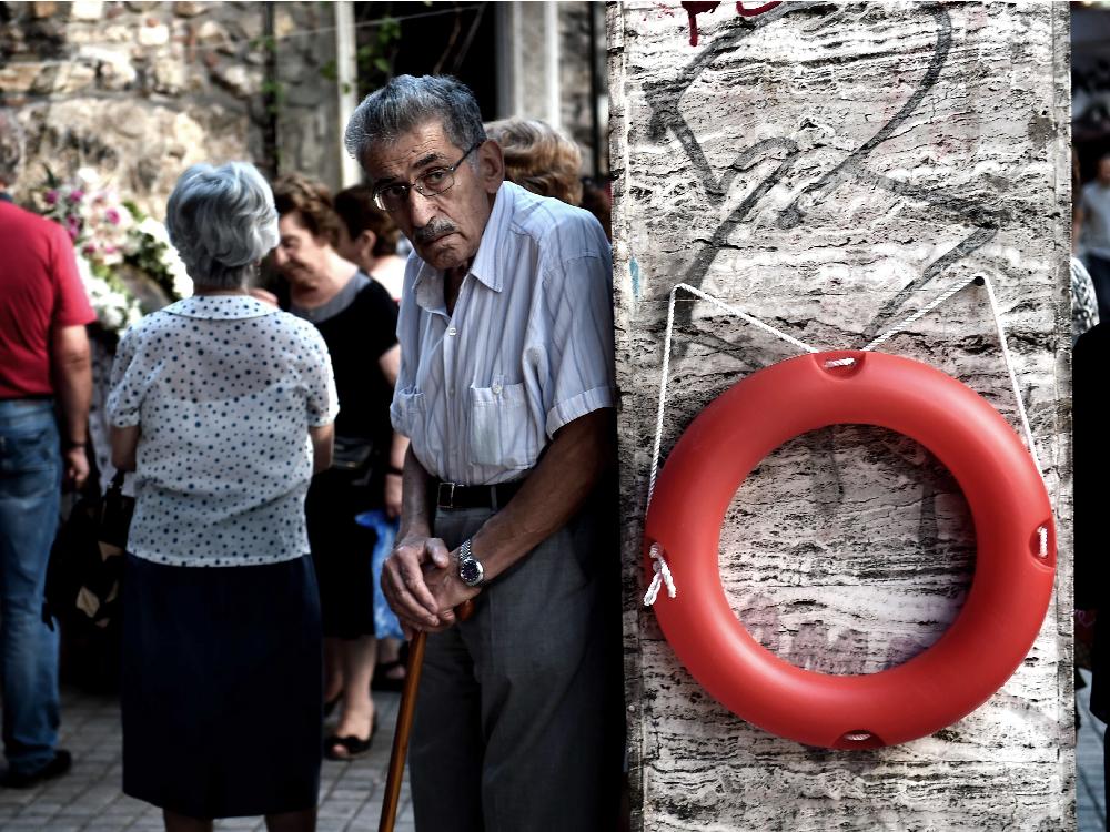 An elderly man stands beside a lifebuoy hanging on a wall outside a chapel in central Athens