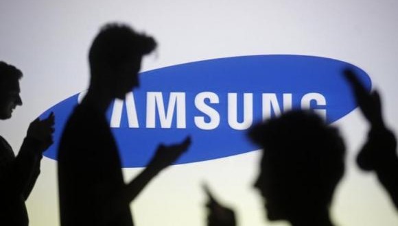 People pose with mobile devices in front of projection of Samsung logo in this