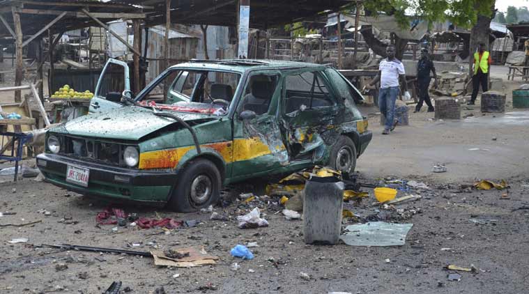 People walk past a damaged car at the site of the bomb explosion in Maiduguri