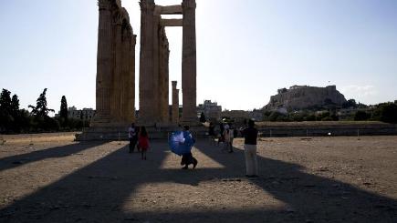 A woman carries a parasol for the strong afternoon sun as a group of tourists walk around the ruined ancient temple of Olympian Zeus with the Acropolis hill in the background in central Athens