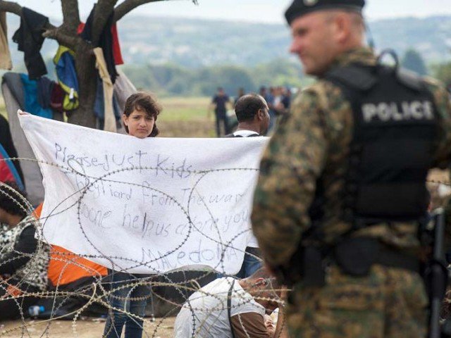 A police officer stand by near a young girl from Syria holding up a towel reading'Please just kids tired from the war no one help us alone you Macedonia as migrants wait behind barbed wire in a noman's land between Greece and Macedonia to cross