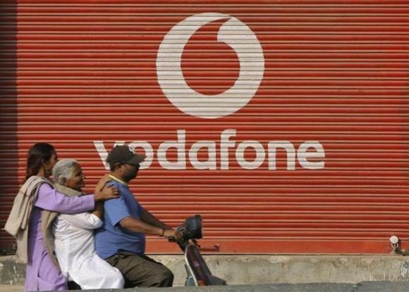 A man and two passengers ride on a scooter past a shop displaying the Vodafone logo on its shutter in Jammu