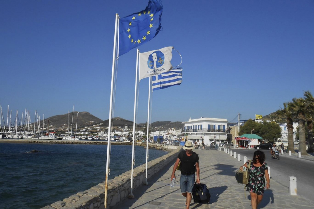 Tourists make their way past a European Union flag and a Greek national flag at the seaside of the island of Paros Greece