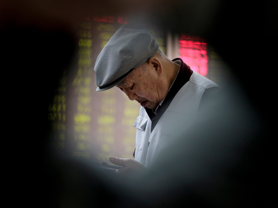 An investor looks at his mobile phone near an electronic board displaying stock prices at a brokerage house in Beijing