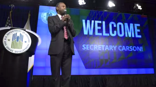 A Black man in a suit and red tie stands before a blue backdrop with the words'Welcome Secretary Carson