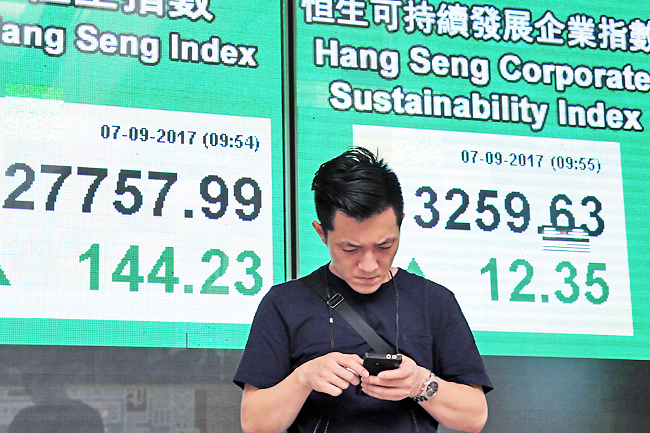 A man uses his smartphone in front of an electronic stock board showing the Hang Seng Index at a bank in Hong Kong