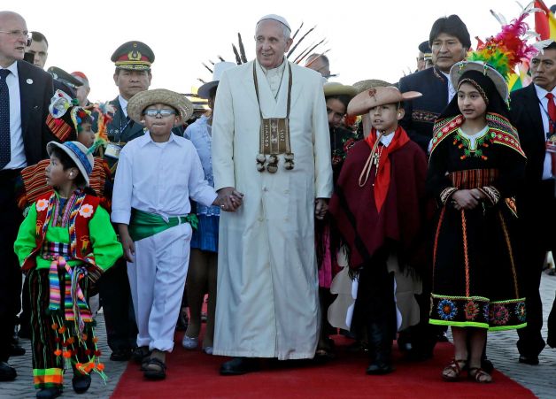 Pope Francis holds hands with children wearing traditional clothing as he walks with Bolivian President Evo Morales background right upon his arrival at the El Alto airport in Bolivia. The pouch Francis