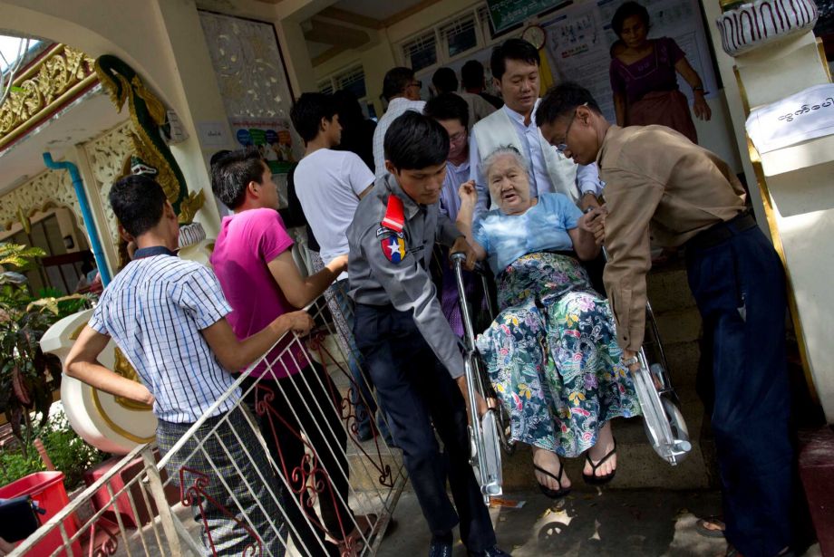 Officials carry an elderly woman in her wheelchair down the stairs of a polling station after casting her vote in Yangon Myanmar Sunday Nov. 8 2015. Myanmar voted Sunday in historic elections that will test whether popular mandate will help loosen the