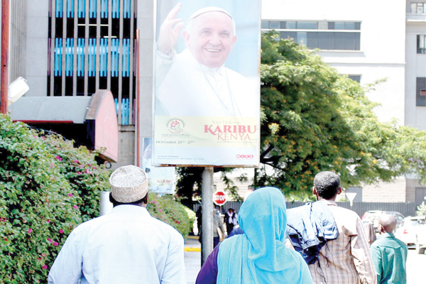 Pedestrians walk past Pope Francis bill board along City Hall Way yesterday as the preparation to welcome him tomorrow are in top gear