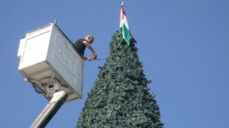 A worker in Bethlehem West Bank wraps festive lights around a tall Christmas tree topped by a Palestinian flag