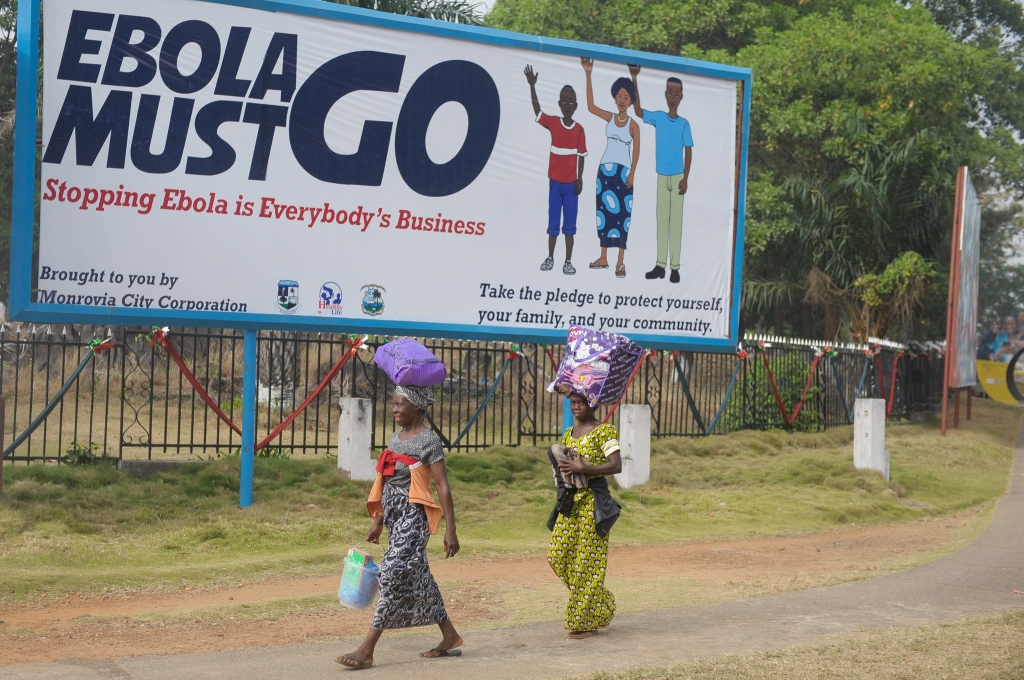 Two women walk in front of a billboard which says'Ebola must go. Stopping Ebola is Everybody's Business in Monrovia Liberia