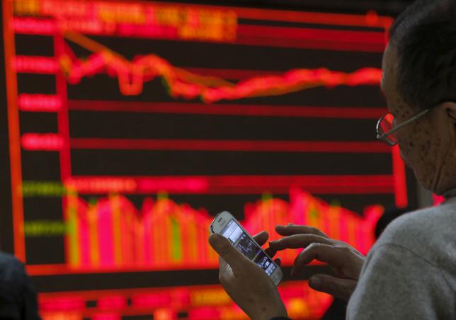 A man checks stock information on his mobile phone in front of an electronic board showing stock information at a brokerage house in Beijing China