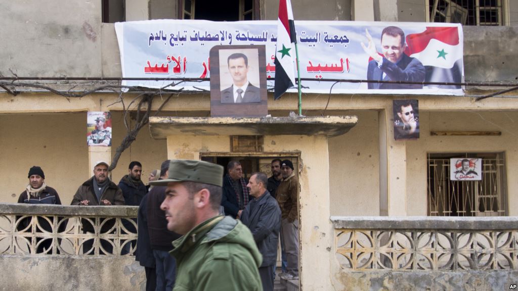 FILE- A Syrian police officer walks past a group of local people standing at a building with a portrait of Syrian President Bashar al Assad in Salma Syria Jan. 22 2016