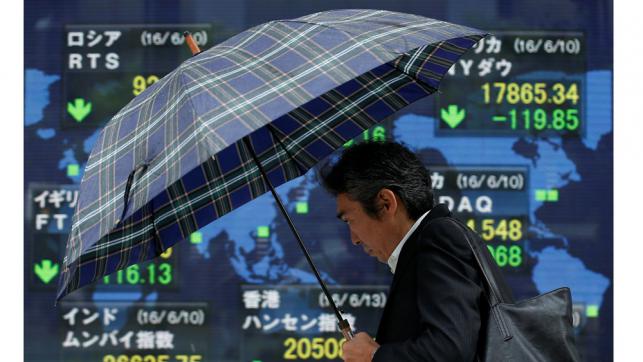 A pedestrian holding an umbrella walks past an electronic board showing the stock market indices of various countries outside a brokerage in Tokyo Japan on Tuesday