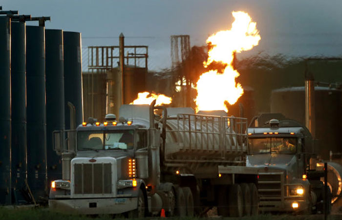 Drivers and their tanker trucks capable of hauling water and hydraulic fracturing liquid line up near a natural gas burn off flame and storage tanks in Williston North Dakota in this file